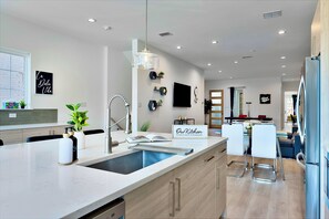 A modern kitchen with a large island, stainless steel appliances, and a dining area with a blue chair. - [Hot Tub] Fun Dallas Downtown House w/ Games+Arcades+Gym+EV Plug - Sleeps 12 (Dallas)