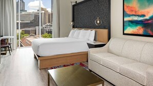 Habitación de hotel moderna con una cama grande, un sofá, una mesita y vistas a la ciudad a través de amplios ventanales.