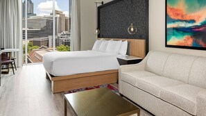 Moderni hotellihuone, jossa on suuri sänky, sohva, pieni pöytä ja suuret ikkunat, joista avautuu näkymä kaupunkiin.