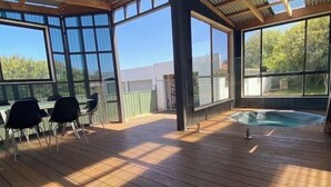A covered outdoor area with a hot tub, wooden deck, and large windows offering views of the surrounding area. - LUXURY HOME  WITH KING BED, HOT TUB, WiFi, AC , Arcade games Netflix sleeps 9  (Portland)