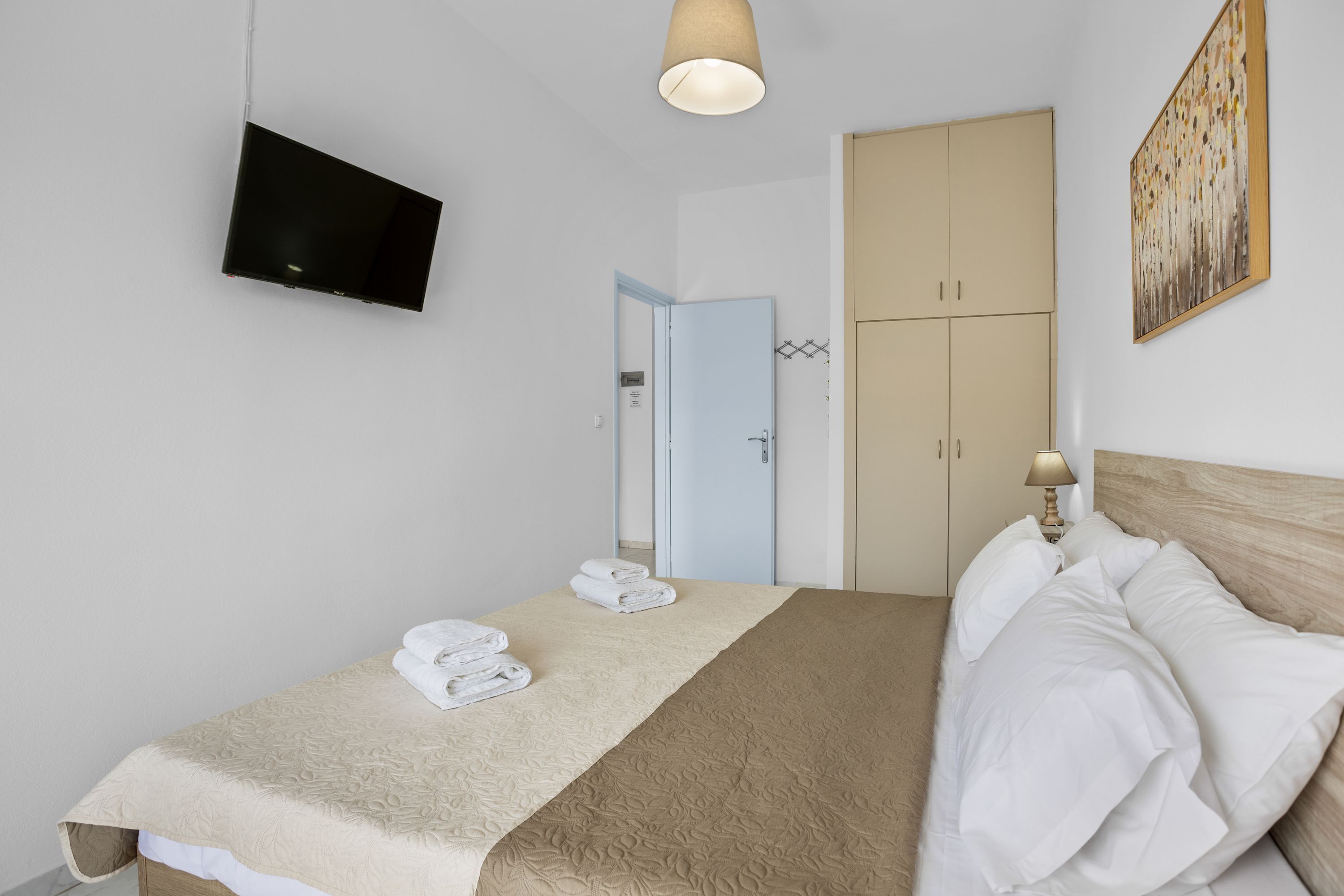 Uma cama bem arrumada com edredom bege e branco, uma TV de tela plana na parede, um painel de cabeceira de madeira e uma porta que dá para outro quarto.