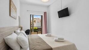 A hotel room with a bed, a TV, a balcony with a view of buildings, and a chair. - Seafront Apartment in Chania 7 (Chania)