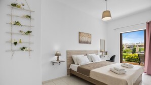A bedroom with a bed, a bedside table, a lamp, a wall-mounted shelf with plants, a painting, and a view of the outside. - Seafront Apartment in Chania 7 (Chania)