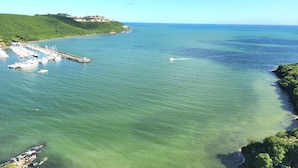 A marina with boats docked, a clear view of the sea, and a green hillside in the background.