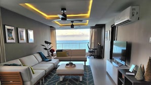 A modern living room with a sofa, coffee table, and TV. There is a balcony with a view of the sea.