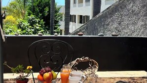 Una mesa con un frutero, vasos de jugo y una jarrón, con palmeras y un edificio al fondo.