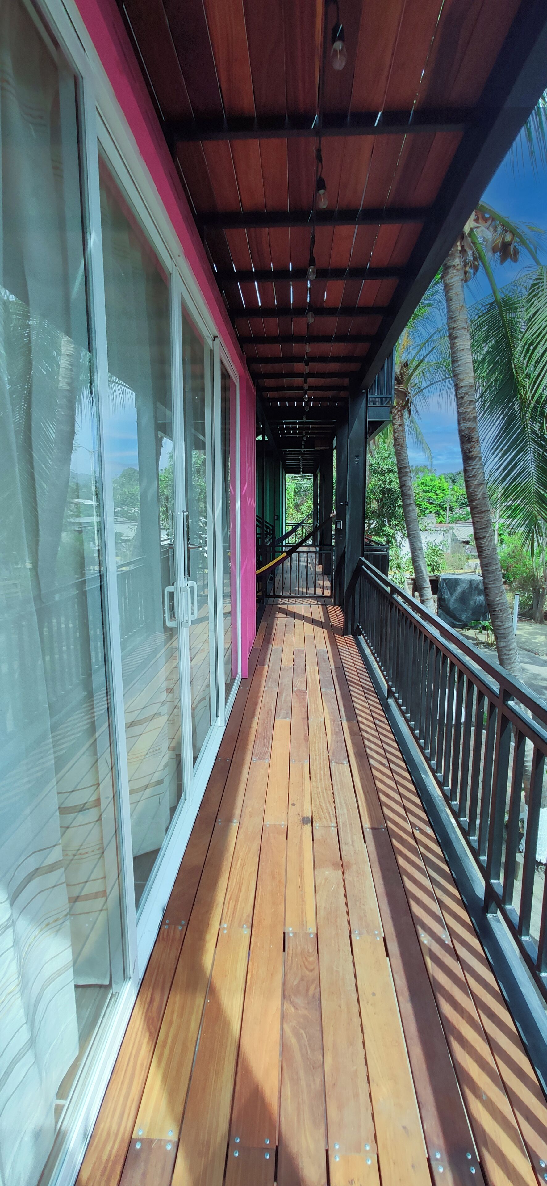 A wooden deck with glass doors leading to a covered area, surrounded by greenery and palm trees.