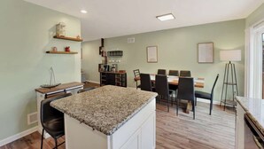 Una cocina con encimera de granito, un comedor con mesa y sillas, y un estante pequeño con objetos decorativos.