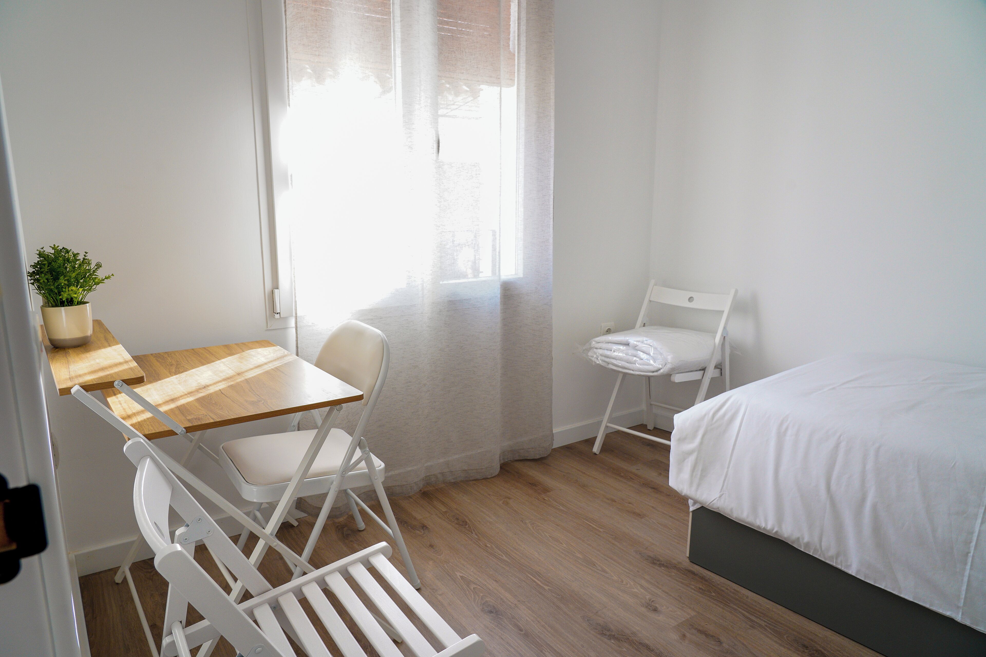 Una habitación con escritorio de madera, silla blanca, cama y una pequeña planta en maceta.
