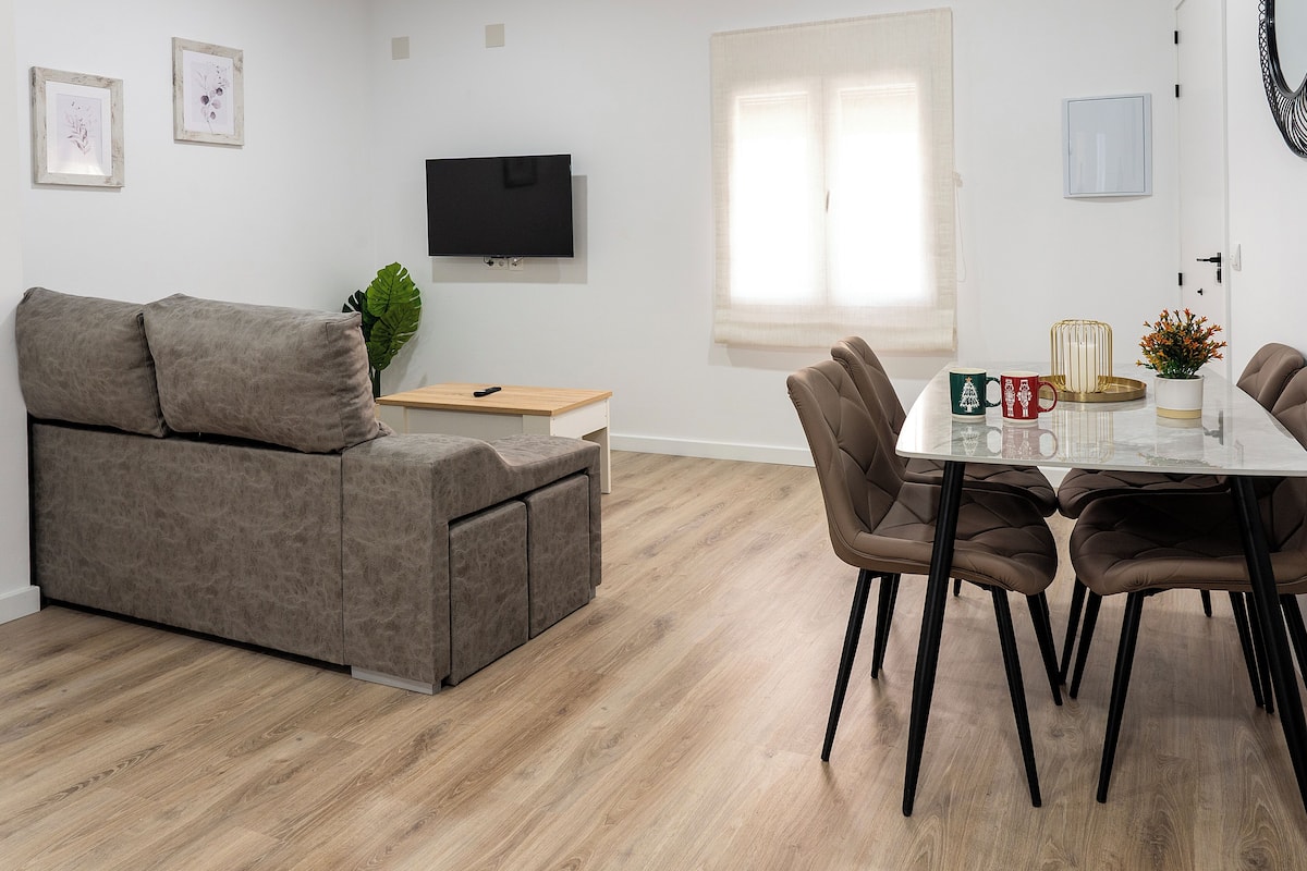 Una sala de estar moderna con un sofá gris, una mesa de centro de madera y un comedor con una mesa de vidrio y sillas marrones.