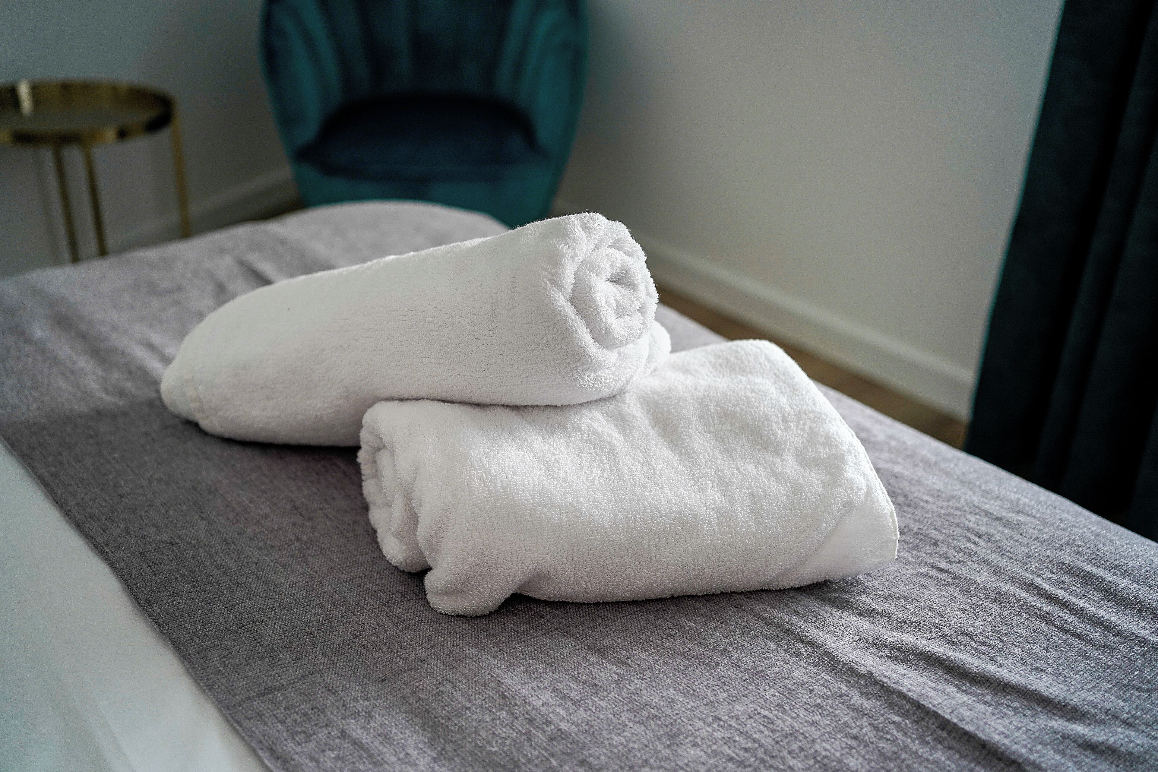Een net opgemaakt bed met twee gevouwen witte handdoeken op een grijze plaid, in een kamer met een blauwgroene stoel en een klein rond tafeltje op de achtergrond.