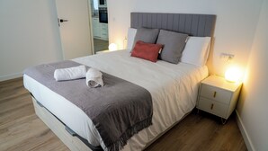 Una cama bien hecha con un cabecero gris, ropa de cama blanca y una manta gris. Hay una mesita de noche blanca con una lámpara y un cajón pequeño. La habitación tiene suelo de madera y una puerta que conduce a un baño.