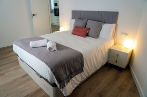 Una cama bien hecha con un cabecero gris, ropa de cama blanca y una manta gris. Hay una mesita de noche blanca con una lámpara y un cajón pequeño. La habitación tiene suelo de madera y una puerta que conduce a un baño.