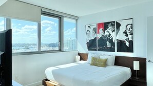 Une chambre d’hôtel moderne avec un grand lit, un téléviseur posé sur un bureau en bois et une vue sur la ville par les fenêtres.