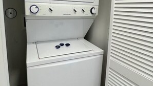 Une petite buanderie blanche, équipée d’un lave-linge et d’un sèche-linge à chargement frontal, d’une petite fenêtre et d’une porte dotée de panneaux à persiennes horizontales.