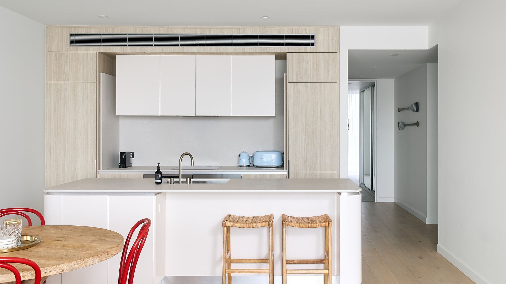 A modern kitchen with white cabinets, a stainless steel sink, and a toaster. There are two wicker stools and a round wooden table with red chairs.