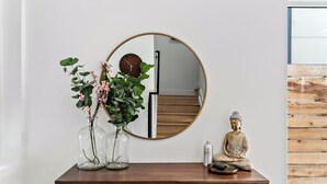 Un tavolo consolle in legno con uno specchio rotondo, due vasi in vetro con piante, una statua di Buddha e una bottiglia di gel disinfettante.
