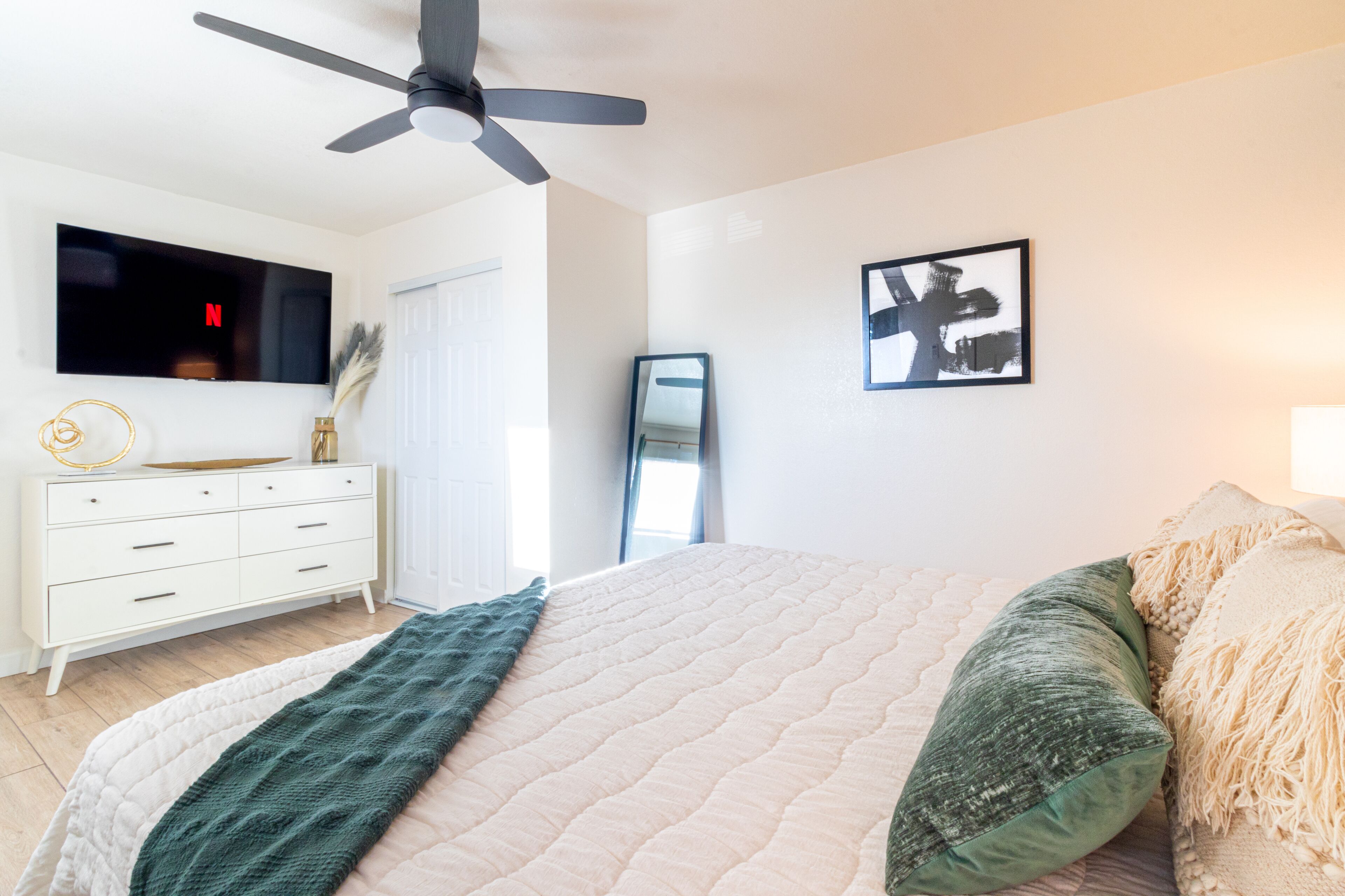 Un dormitorio con cama, ventilador de techo, televisor, cómoda y un cuadro en la pared.