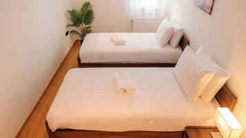 Two single beds with white linens and rolled towels, a bedside table with a lamp, a potted plant, and a window with sheer curtains.