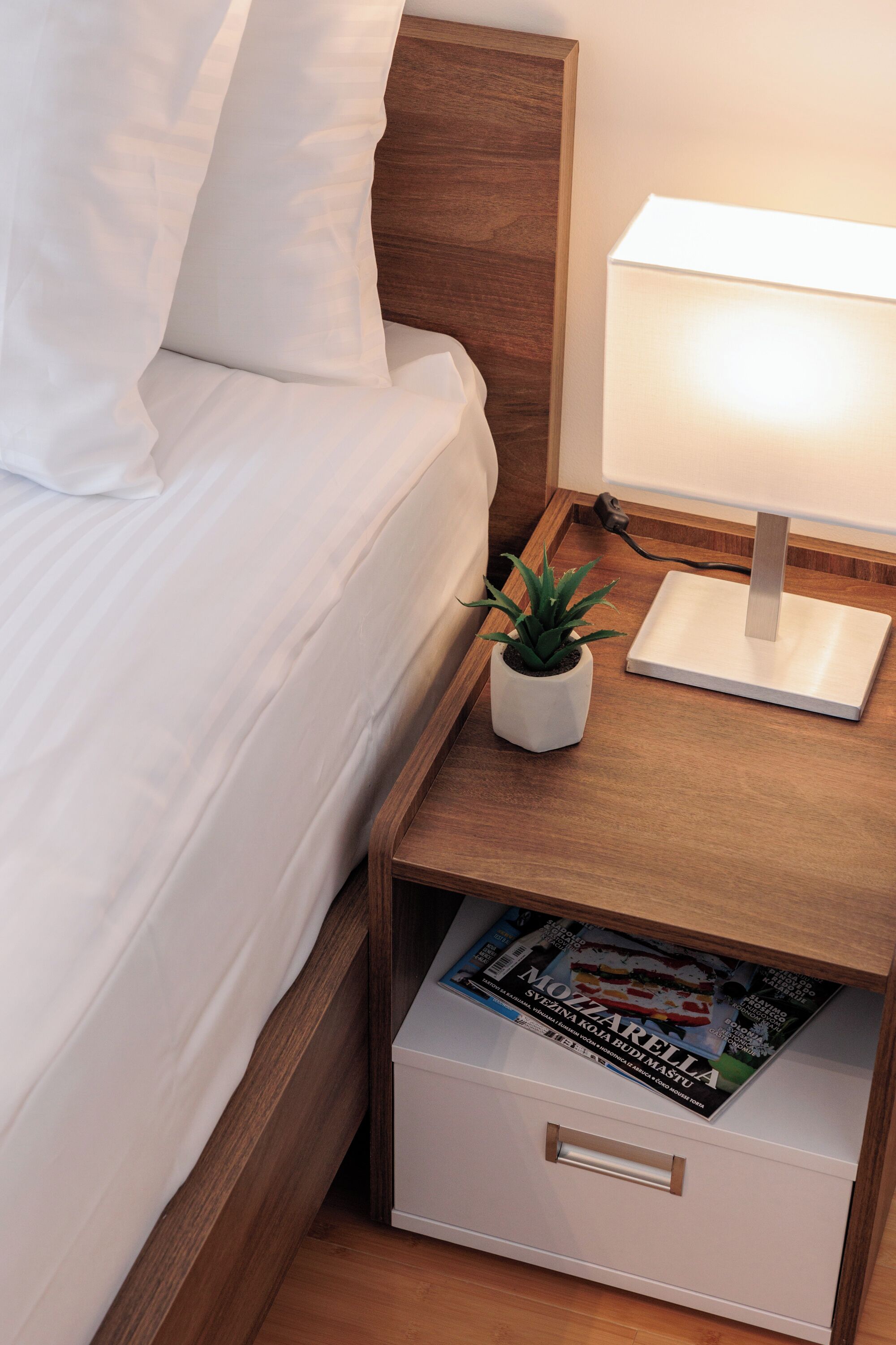 Una cama bien hecha con sábanas blancas, una lámpara de noche, una pequeña planta en maceta y una revista en un cajón.