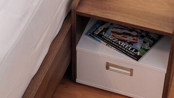 Una cama bien hecha con sábanas blancas, una lámpara de noche, una pequeña planta en maceta y una revista en un cajón.