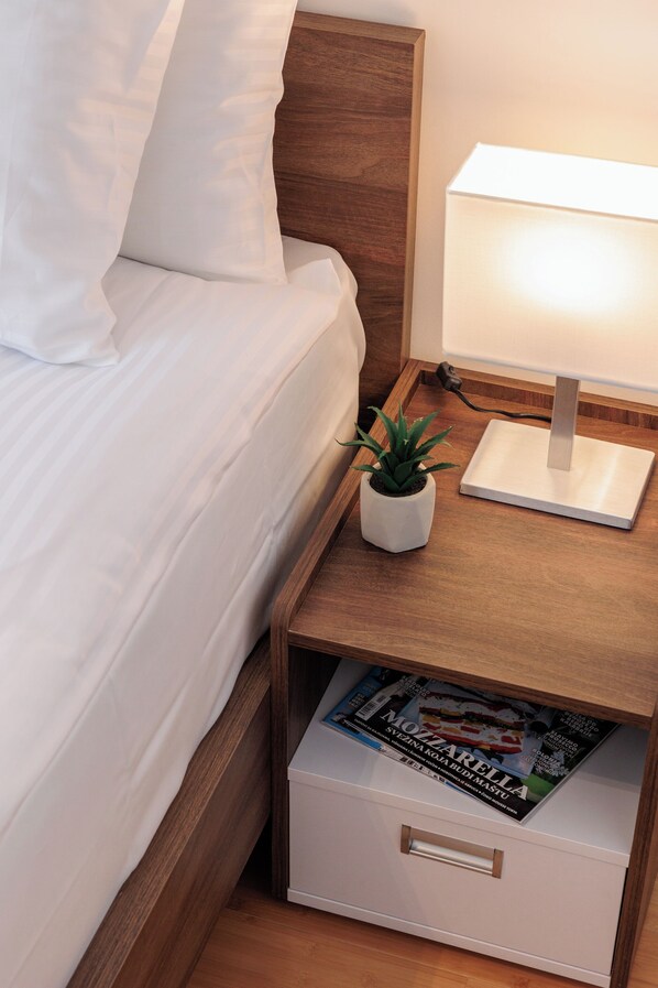 A neatly made bed with white linens, a bedside lamp, a small potted plant, and a magazine in a drawer.