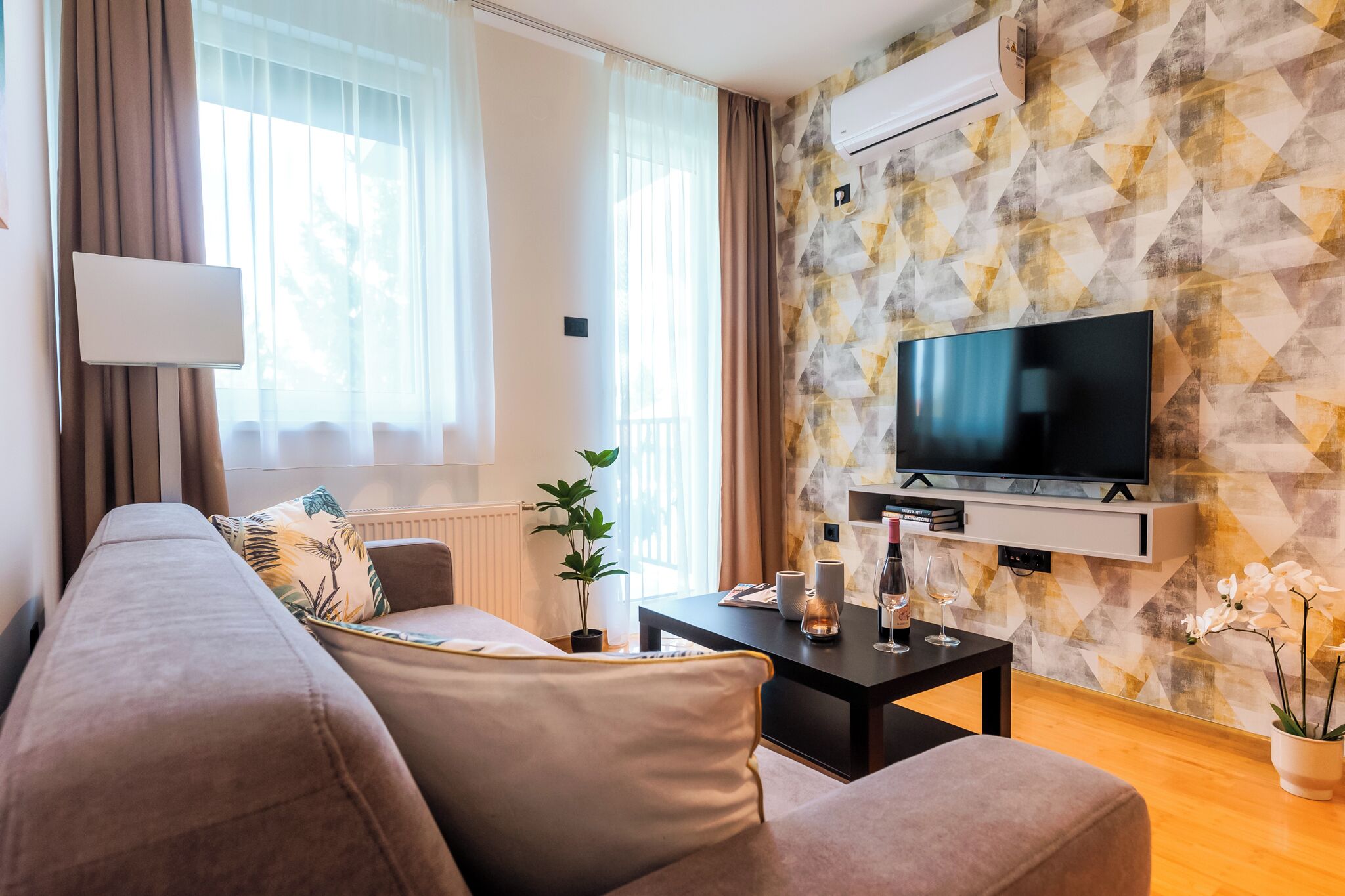 A modern living room with a sofa, a coffee table, a flat-screen TV mounted on a geometric-patterned wall, and a potted plant.