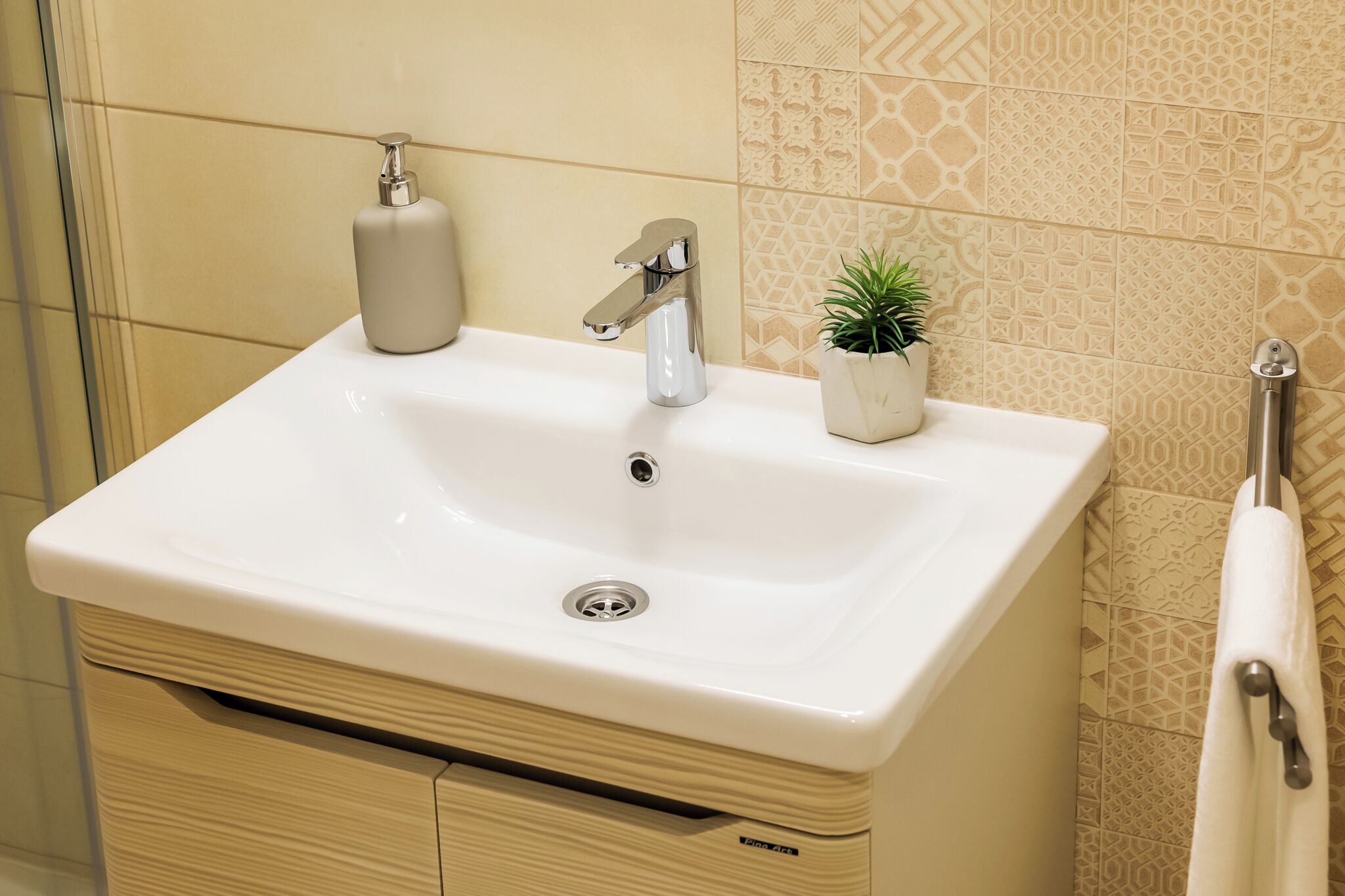 A bathroom with a white sink, a silver faucet, a soap dispenser, a small potted plant, and a towel hanging on a metal rack.