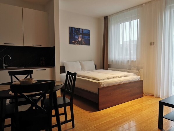 A modern bedroom with a bed, a dining table with chairs, a kitchen area, and a large window with sheer curtains.