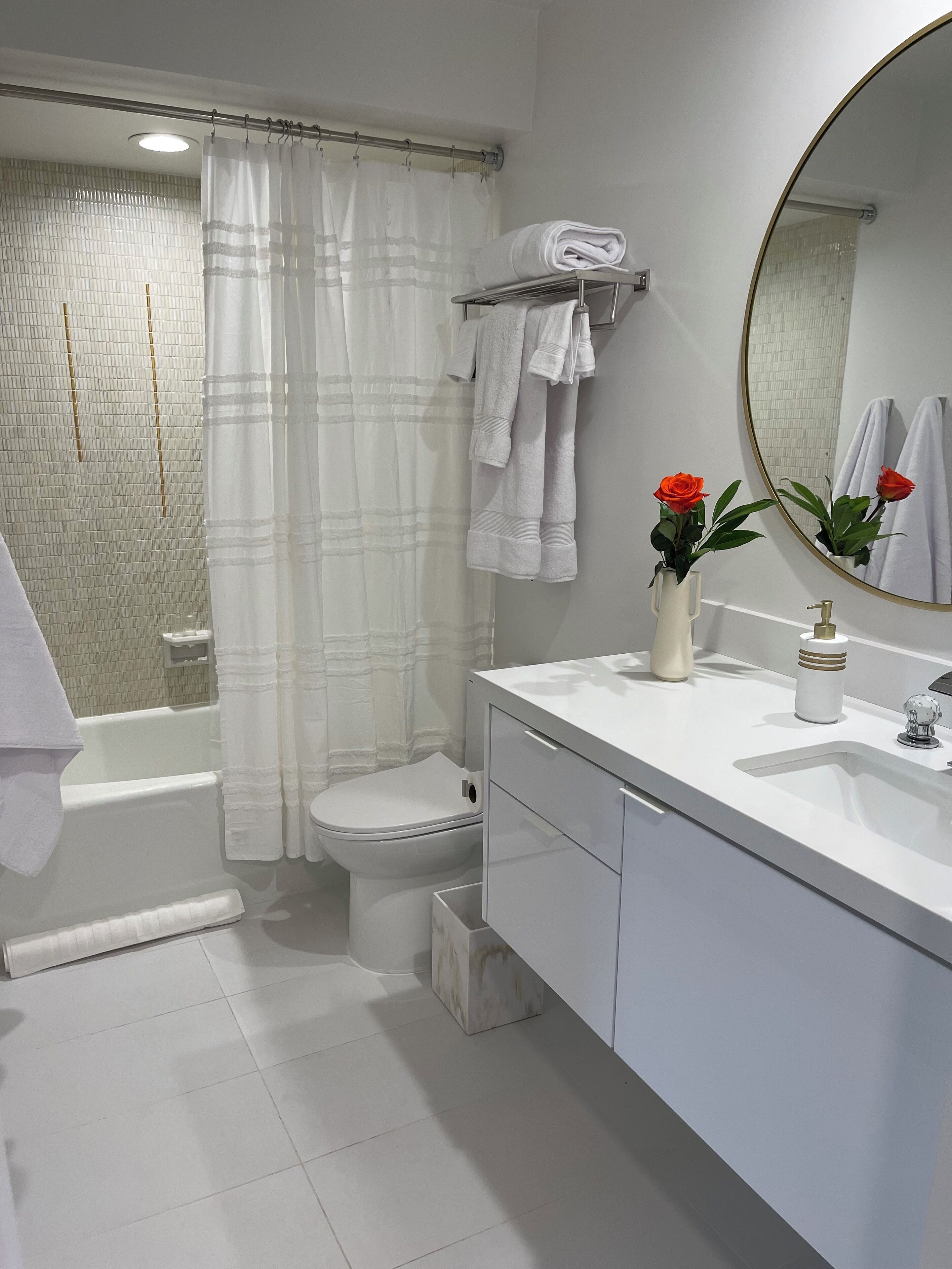 Et badeværelse med et hvidt badekar, toilet og vask. To håndklæder hænger på en bøjle, og der er en vase med en rød rose på vaskeskabet.