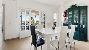 Un comedor con una mesa y sillas blancas, un armario verde con puertas de cristal, y vista a un patio exterior por una puerta abierta.