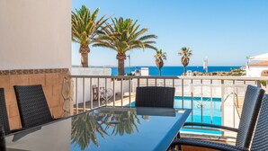 Un balcón con una mesa y sillas de vidrio, con vistas a una piscina y palmeras, y una vista al mar.