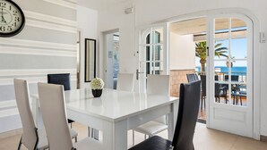 Un comedor con una mesa y sillas blancas, un reloj en la pared y vista al océano a través de un ventanal abierto.
