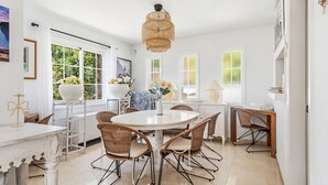 Et lyst køkken med hvide skabe, et rundt spisebord, fletstole og en hængende lampe.