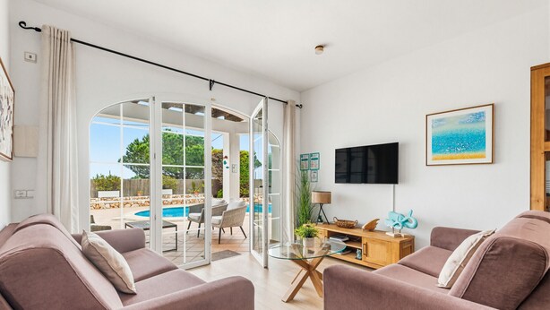 A living room with a sofa, a TV, a glass table, and a view of an outdoor pool area. - Villa Rouss by Escape Home (Calan Porter)