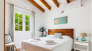 A bedroom with a wooden headboard, a bed with white linens, a nightstand with a lamp, and a window with curtains. - Villa Rouss by Escape Home (Calan Porter)