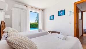A hotel room with two beds, a window with a view of greenery, a wooden headboard, and a wall-mounted air conditioner. - Villa Ana by Escape Home (Santo Tomas)