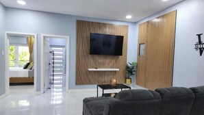 A modern living room with a flat-screen TV mounted on a wooden panel wall, a grey sofa, and a small table with a lamp. - Williams Manor [Across From Ricks Cafe Negril] (Negril)