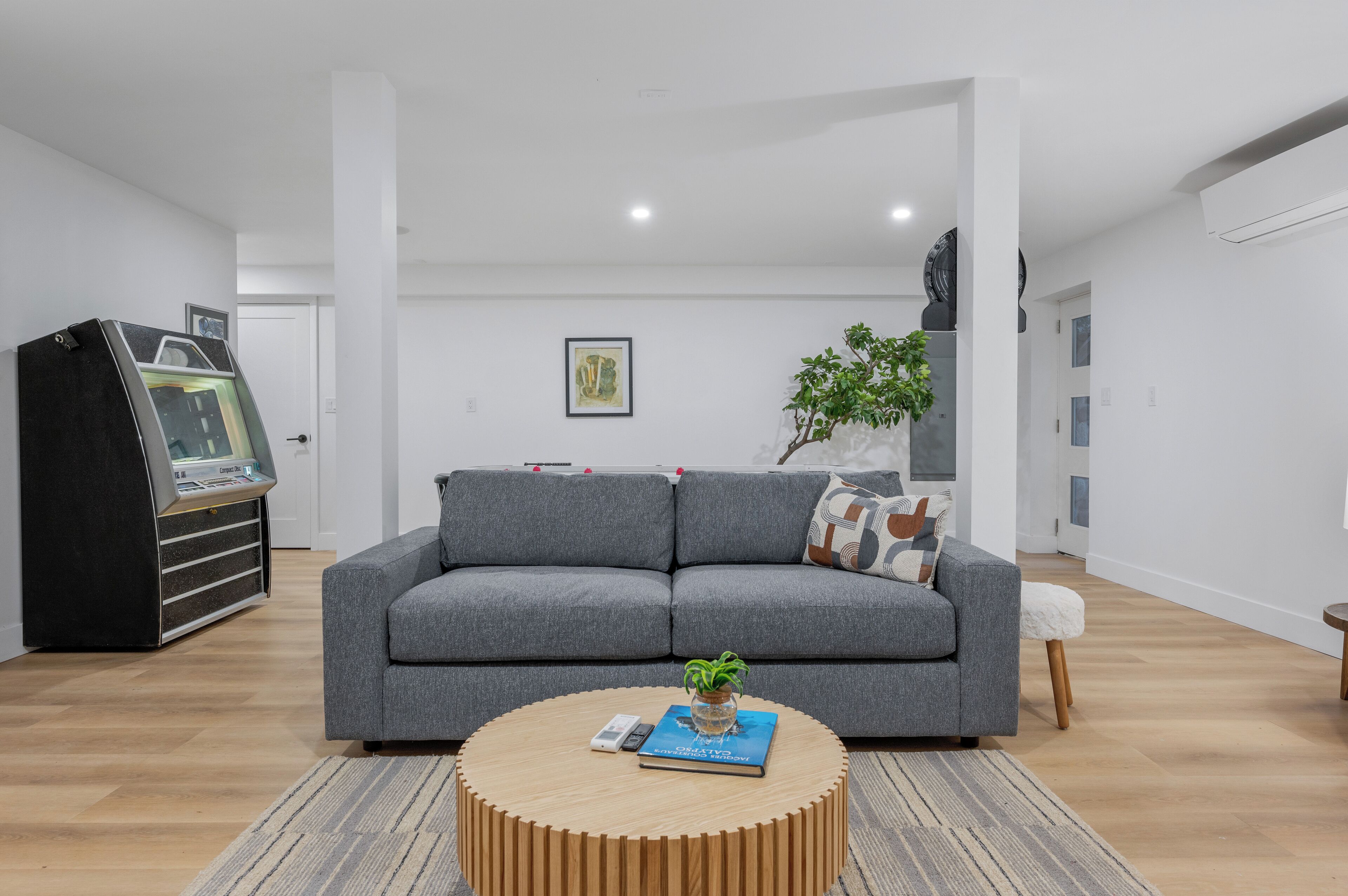 A modern living room with a grey sofa, a round wooden coffee table with a book and a plant, and a gaming console.