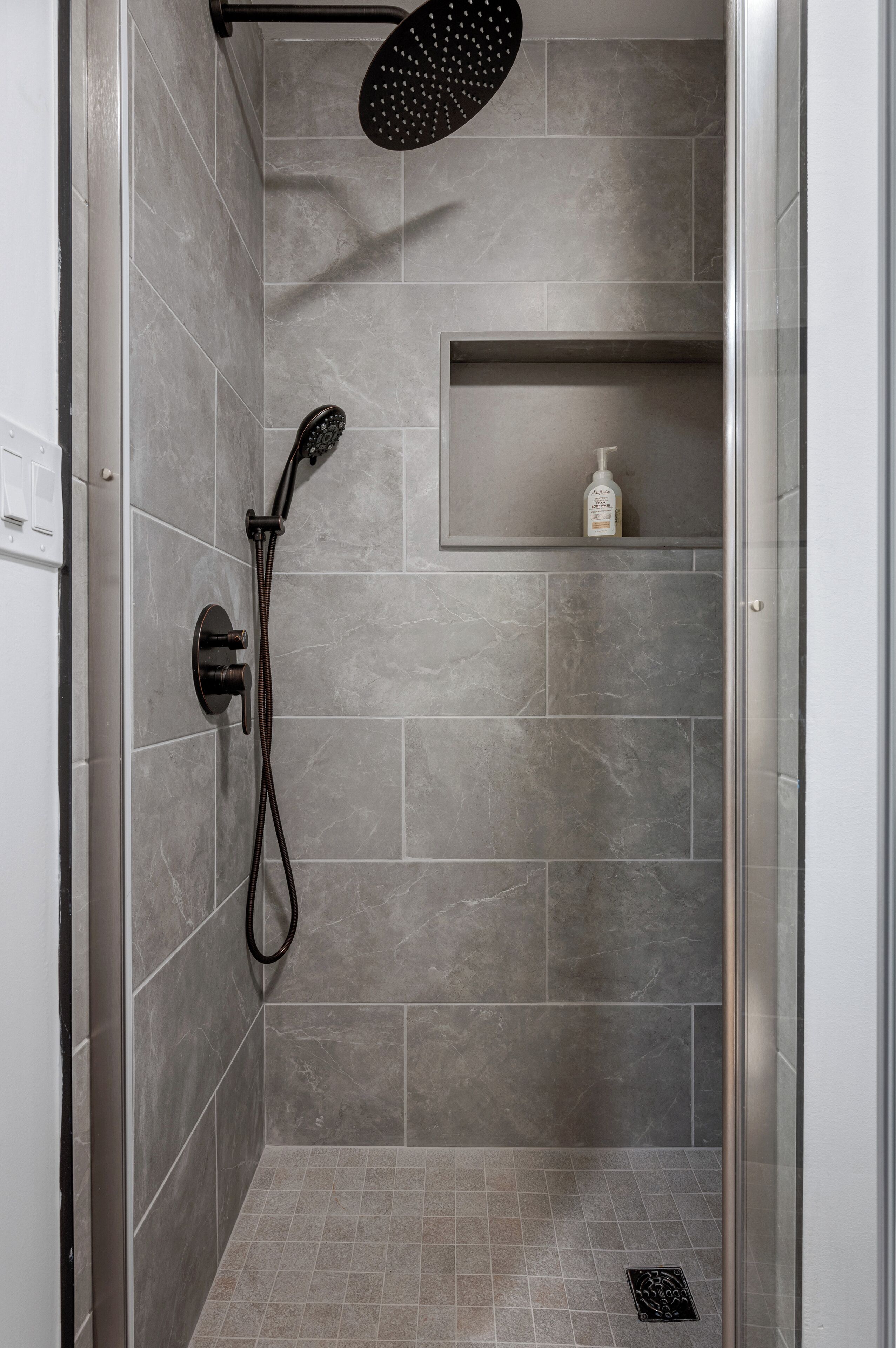 A shower with a handheld showerhead and a handheld hose, grey tiled walls, and a shelf with a bottle.