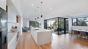 A modern kitchen with a central island, a dining area with a glass partition, and a view of the outdoors. - Austral House - Ultimate Coastal Retreat (Avoca Beach)