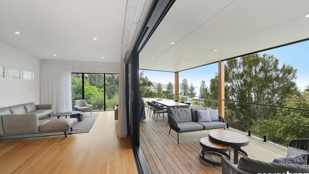 A modern living space with a balcony, wooden flooring, and a view of trees. - Austral House - Ultimate Coastal Retreat (Avoca Beach)