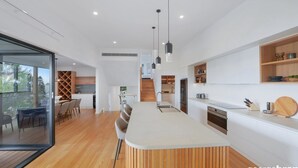 A modern kitchen with a central island, wooden flooring, and built-in shelving. - Austral House - Ultimate Coastal Retreat (Avoca Beach)