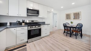 A modern kitchen with white cabinets, a stove, and a microwave. There is a dining table with chairs and a small plant. - Dapper 2-Bedroom Abode: Chic & Cozy (Sacramento)