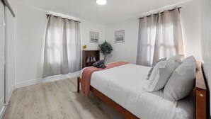 A bedroom with a bed, wooden floor, curtains, a bedside table, a plant, and two framed pictures on the wall. - Dapper 2-Bedroom Abode: Chic & Cozy (Sacramento)