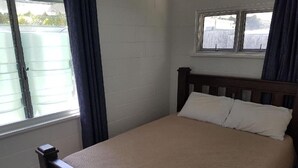 Une chambre avec un lit en bois, des murs blancs, une fenêtre avec des rideaux et une vue sur un espace clos à l’extérieur.