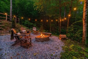 A forested outdoor seating area with a fire pit, wooden chairs, and string lights. - Rushing Creek Retreat, Serene Cabin with Great Views! (Sylva)