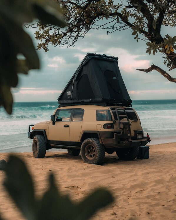 A beige off-road vehicle with a black tent on a sandy beach, overlooking the ocean.