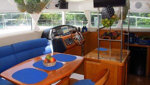 A boat's interior with a blue sofa, wooden table with blue placemats, a steering wheel, and a TV mounted on a stand. -  Tropical Yacht Charters. - Family Vacation (Placencia)
