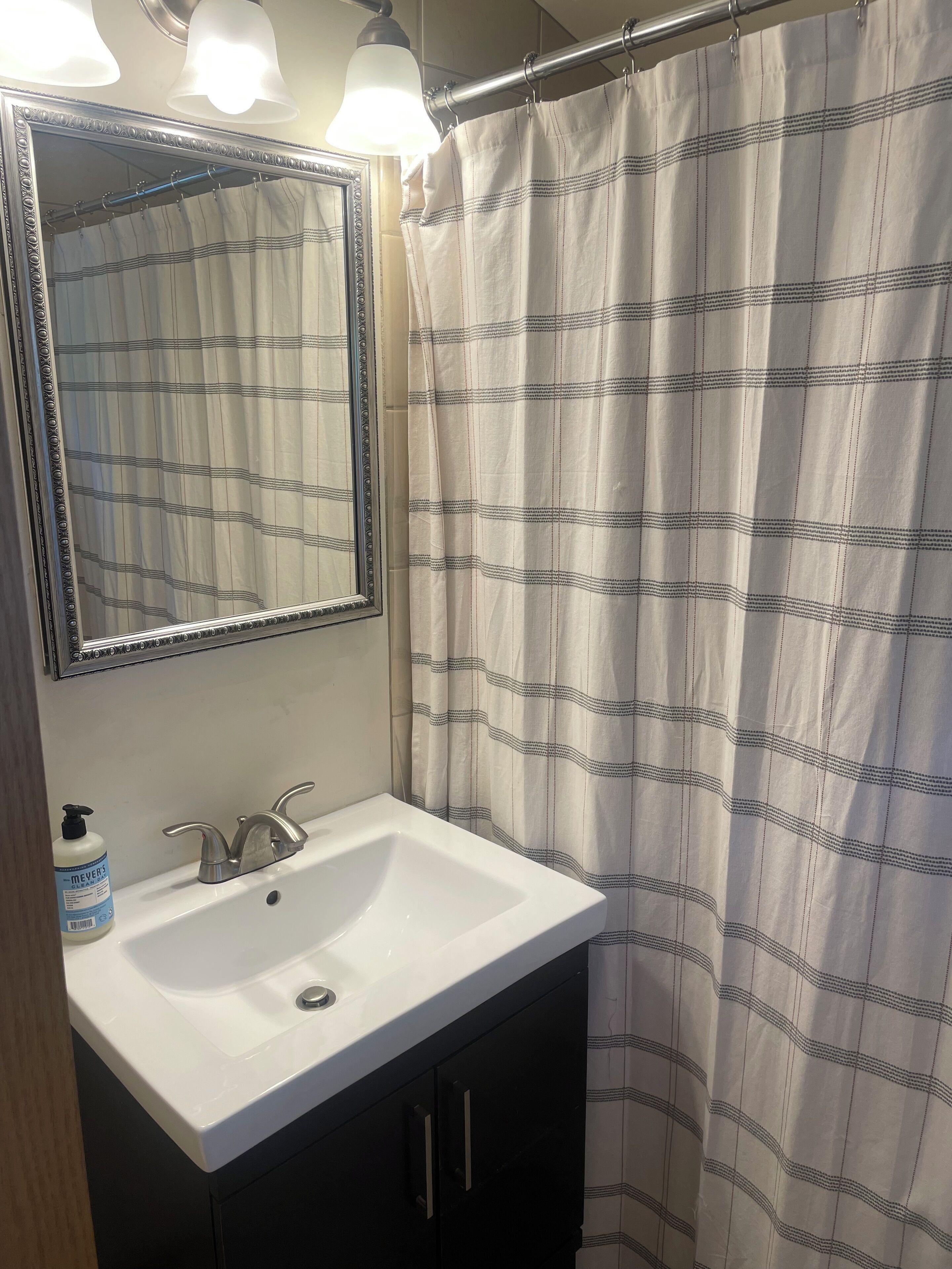 Ein Badezimmer mit weißem Waschbecken, einem Spiegel mit Zierrahmen und einer Duschvorhang mit Karomuster.
